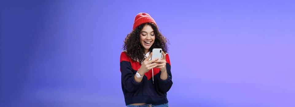 Lifestyle. Joyful and sociable stylish caucasian woman with curls in warm beanie having fun laughing and chuckling as using smartphone typing funny message looking at mobile phone screen happily over blue wall.