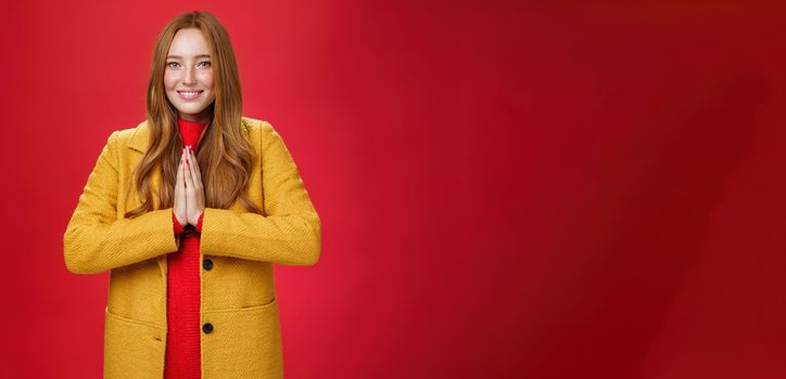 Girl welcomes us with buddhist namaste gesture pressing palms together over chest smiling friendly and delighted with carefree emotions holding hands in pray, posing happily over red background.