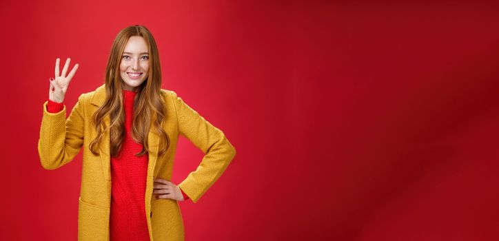 Cute friendly-looking ginger girl showing three reasons buy product smiling broadly making third reservation, looking delighted and confident in her choice posing against red background in yellow coat.