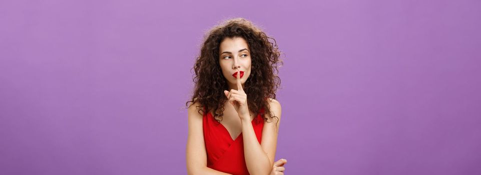 Keep it secret babe. Confident and tricky caucasian female with curly hairstyle in red elegant dress shushing looking left curious wanting tell secret showing shh gesture with index finger over mouth.