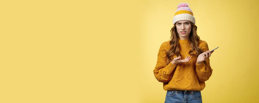Confused cringing annoyed young woman shocked how friend talked her frowning raise hand dismay look frustrated camera holding smartphone finish video call unpleasant note, yellow background.