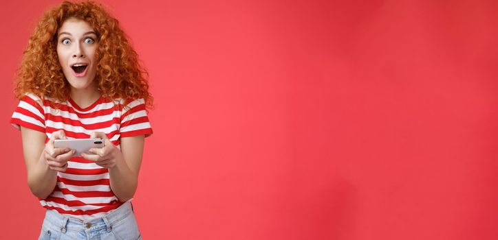 Lifestyle. Impressed excited good-looking redhead curly woman hold smartphone horizontal open mouth fascinated stare camera thrilled astonished win passed level game happy playing cool app red background.