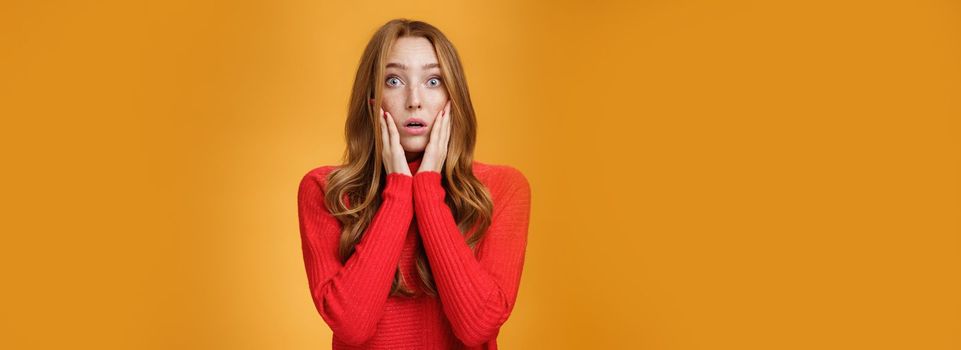 Worried shocked redhead female reacting to stunning troublesome news holding palms on cheeks open mouth and raise eyebrows astonished, posing concerned against orange background. Copy space