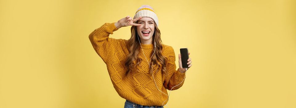 Positive carefree charming young girl show peace gesture wearing wired earphones showing smartphone screen promoting app cool brand new mobile phone, laughing carefree yellow background.