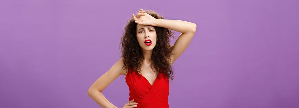 Woman fainting feeling bad whiping sweat of forehead standing drained and exhausted over purple background in red stylish dress expressing gloomy and unhappy feelings wanting some help. Copy space