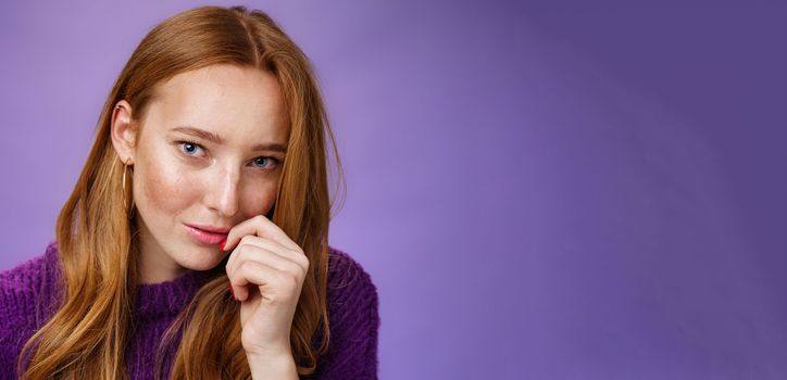 Ginger girl charmes with sensual romantic gaze. Portrait of confident flirting and attractive redhead female tilting head touching lips and looking daring at camera against purple background.