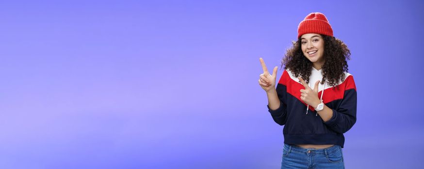 Lifestyle. Portrait of friendly good-looking woman with curly hair in red warm beanie pointing at upper left corner and smiling delighted as promoting cool copy space against blue background.