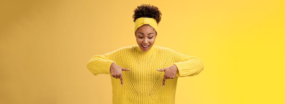 Impressed enthusiastic smiling fascianted african-american woman in sweater headband look pointing down index fingers gasping astonished fascinated cool thrilling promo, standing yellow background.