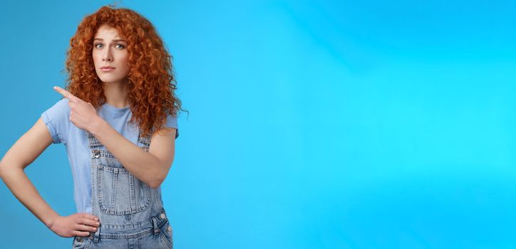 Lifestyle. Unsure silly timid hesitant cute redhead curly-haired ginger girl frowning uncertain asking your opinion questioned look camera pointing upper left corner confused worried blue background.