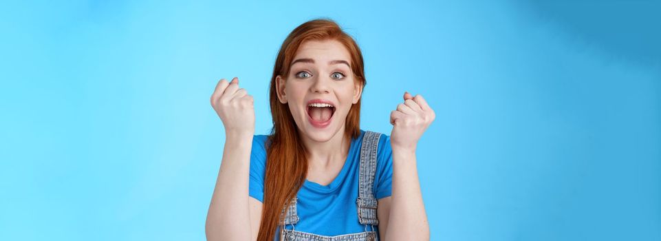 Close-up cheerful lucky redhead woman win lottery, fist pump yell yeah hooray, winning bet, smiling gladly rejoicing excellent news, celebrating success, triumphing happily, stand blue background.