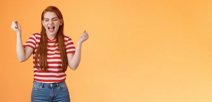 Luck cute redhead girl, make fist pump cheerfully yell yes, close eyes, winning lottery, feeling amazed, celebrate success and victory, stand orange background, triumphing, rejoicing good news.