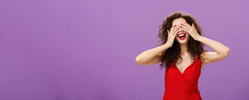 Wife waiting for husband present gift for anniversary closing eyes with palms and smiling broadly peeking through fingers being charmed and excited standing in seductive red dress over purple wall. Emotions concept