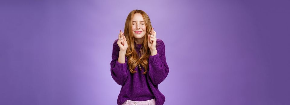 Girl makes wish eager dream come true. Atractive hopeful and positive young redhead woman in purple sweater close eyes and smiling as cross fingers for good luck, praying and anticipating miracle.
