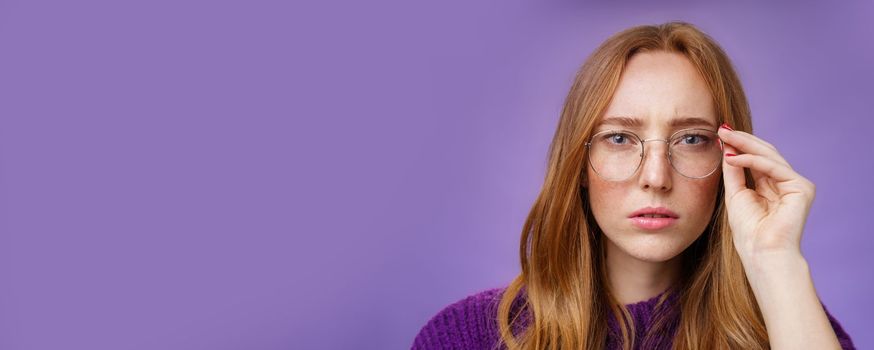 Unsure and serious-looking focused redhead woman frowning and squinting as looking in prescribed glasses at camera, touching frame reading sign, solving puzzle over purple background.