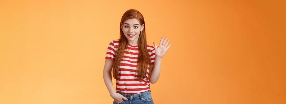 Hello how are you. Cute modest redhead newbie student female say hi, waving hand friendly, get know coworkers, smiling joyfully, introduce herself, welcome home, stand orange background.