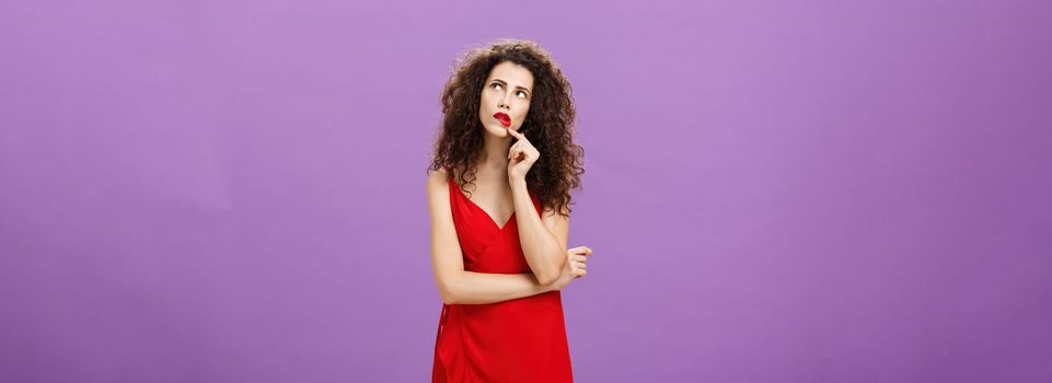 Elegant thoughtful woman. in trendy red dress with curly hairstyle and party makeup touching lip looking at upper right corner thinking how make impression during conversation over purple background.