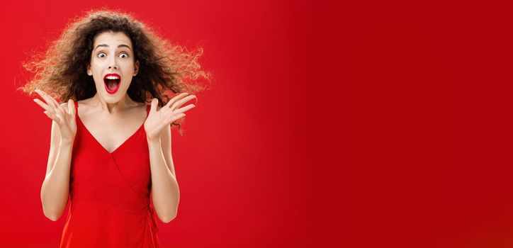 Portrait of surprised and astonished speechless curly-haired woman. in red luxurious dress dropping jaw from amazement popping eyes at camera and gesturing emotive and shocked over background.