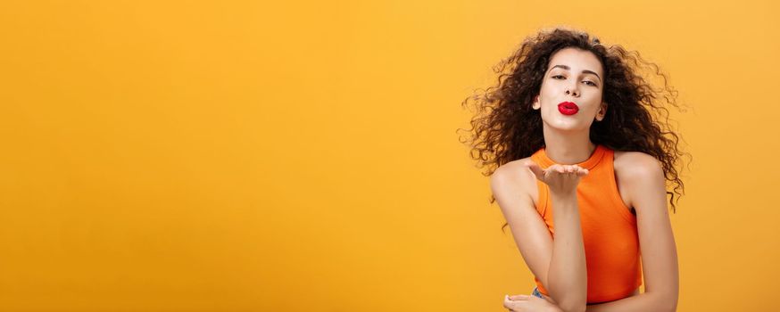 Flirty confident and sensual stylish woman with curly hairstyle and red lipstick folding lips holding palm near mouth sending kiss while hair flicks on air being in romantic mood over orange wall. Body language concept
