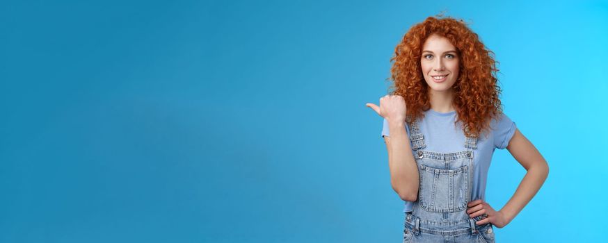 Lifestyle. Cheerful curious lively redhead good-looking curly-haired girl wearing summer overalls t-shirt pointing left thumb look camera intrigued excited explore new stores friends walking shopping mall.