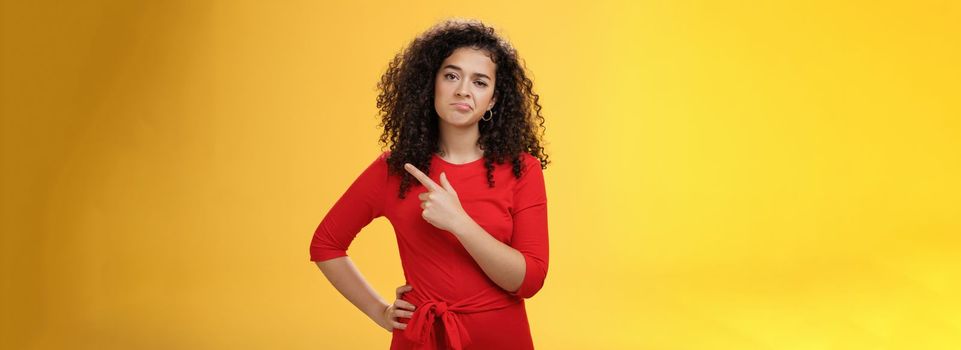 Portrait of unimpressed careless and bored young curly-haired female coworker in dress making upset smile pointing at upper right corner looking with indifference and disappointment. Body language concept