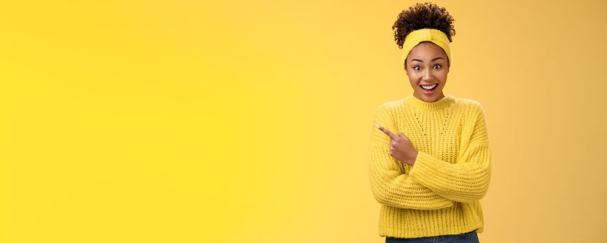 Amused charismatic smiling black cute girl. in headband sweater widen eyes drop jaw astonished hear about awesome interesting new place talking friend standing yellow background pointing left amazed.
