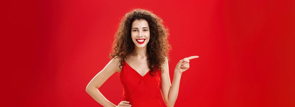 Girl suggesting ask her boyfriend. Charming elegant and stylish young female with curly hairstyle in red evening dress holding hand on hip and pointing left while showing friend where buy clothes.