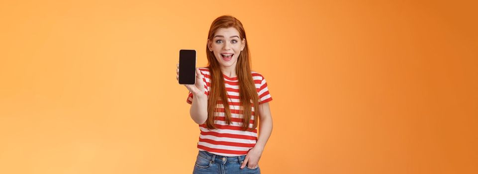 Excited cute outgoing redhead female impressed showing app, hold smartphone introduce gadget feature, smiling surprised, look enthusiastic like application, brag game score, orange background.