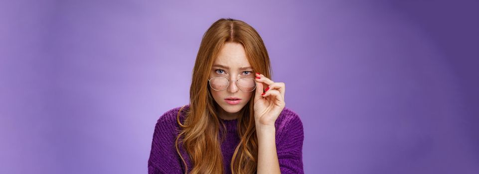 Lifestyle. Questioned and unsure cute smart female detective with ginger hair looking suspicious from under forehead taking off glasses and looking at camera doubtful and uncertain over purple background.