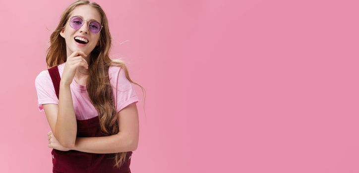 Self-confident good-looking pretty young lady with wavy hair floating on air raising head up having high self-esteem touching chin laughing and smiling wearing trendy sunglasses and dungarees. Body language and emotions concept