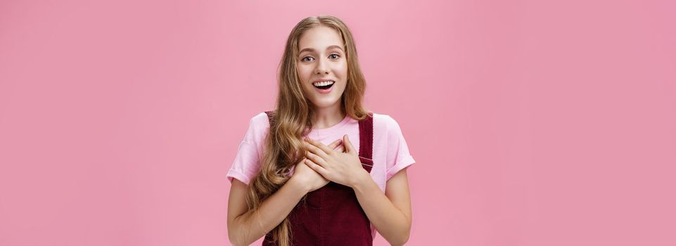 Portrait of thankful and pleased happy young girl receiving surprise gift on b-day pressing palms to hear in gratefyl gesture open mouth from amazement and joy thanking over pink background. Body language and emotions concept