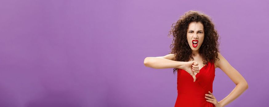 Bullshit I hate your lame ideas. Displeased and pissed lady boss with curly hairstyle in red evening dress growning grimacing from hate and anger showing thumbs down in rejection and disapproval. Copy space