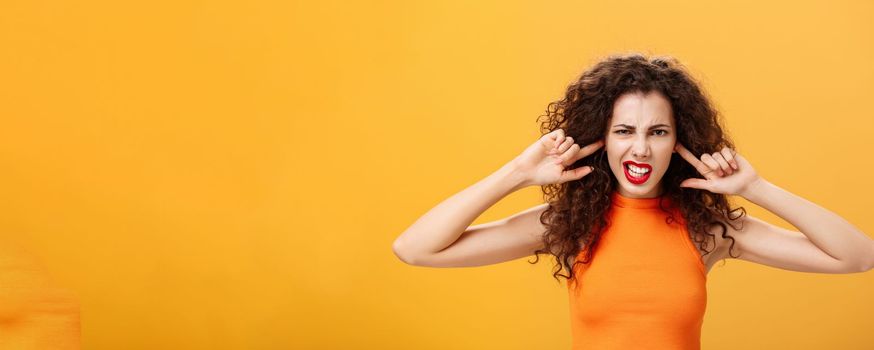 Waist-up shot of irritated bothered stylish woman. with curly hair and red lipstick squinting clenching teeth waiting for loud bang covering ears with index fingers being annoyed of noisy neighbours.