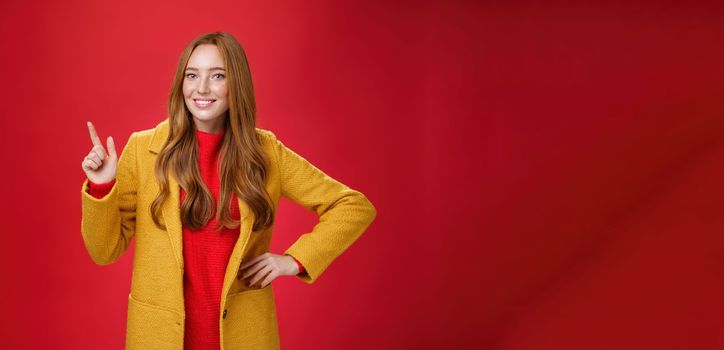 Charming young female in yellow coat holding hand on waist and pointing at upper left corner, smiling broadly as showing or promoting interesting proposal over red background, in good mood.