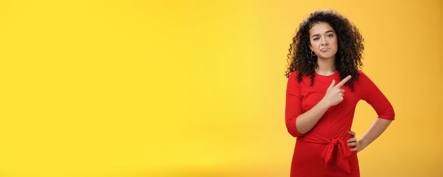 Portrait of unimpressed careless and bored young curly-haired female coworker in dress making upset smile pointing at upper right corner looking with indifference and disappointment. Body language concept