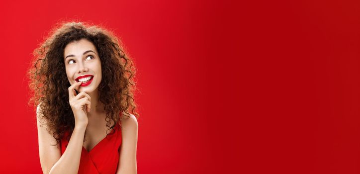 Cute charismatic and tender dreamy woman with curly hairstyle in red dress biting finger and smiling with desire in eyes looking at upper right corner daydreaming picturing object she wants. Copy space