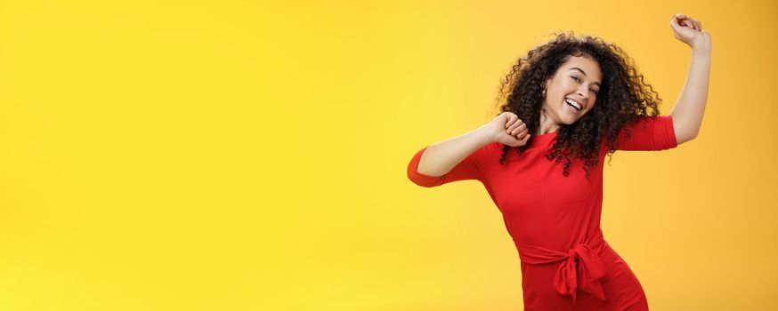 Carefree girl feeling uplifted and joyful dancing in red dress raising hands up happily tilting head and smiling broadly at camera as enjoying vacation, celebrating holidays over yellow background.