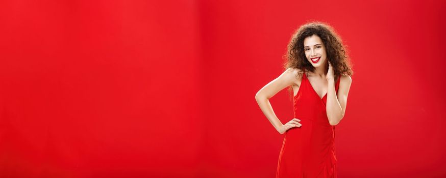 Woman posing in front of boyfriend in new stylish dress ready go out for dinner in romantic restaurant touching neck gently holding hand on hip standing in flirty and feminine pose over red background. Copy space