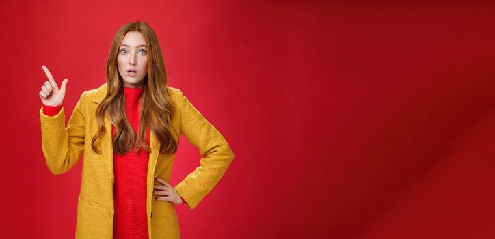 Shocked and stunned cute, confused ginger girl in yellow coat feeling worried and surprised open mouth lifting eyebrows amazed pointing at upper left corner, posing against red background.
