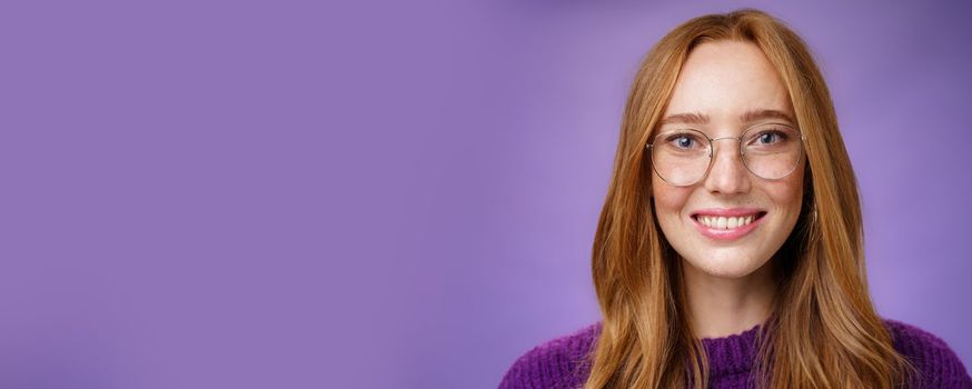 Headshot of smart good-looking joyful and friendly redhead 20s female in transparent glasses smiling broadly and looking hopeful, optimistic at camera feeling happy against purple background.