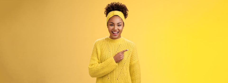 Friendly good-looking enthusiastic black charming girl in headband sweater smiling broadly amused pointing left awesome place showing you perfect blank space advertisement, posing yellow background.