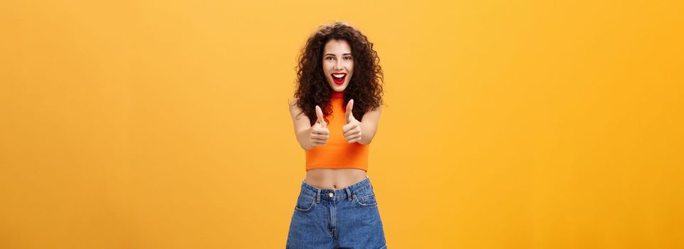 Charismatic ambitious and outgoing charming caucasian. woman with curly hairstyle and red lipstick showing thumbs up gesture in like or approval smiling joyfully being supportive over orange wall.