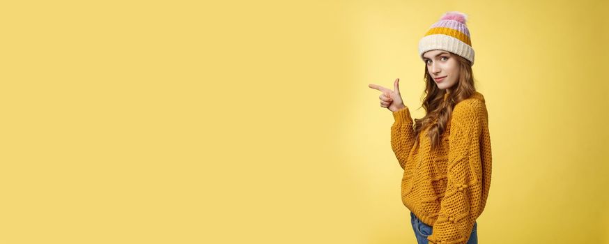 Profile shot confident flirty fashionable young 20s girl turning camera smiling pointing left inviting go together showing curious promotion cool place hang out, standing yellow background relaxed.