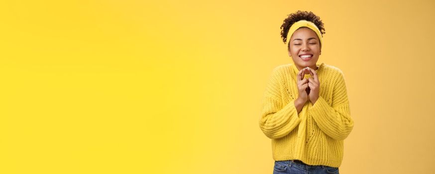 Charming optimistic carefree young smiling african-american woman. close eyes praying hold palms together happily joyfully wishing dream come true feel lucky day standing cheerful yellow background.