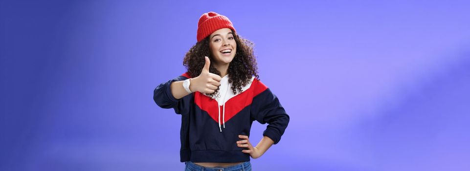 Nice work, keep up. Delighted and supportive charming sportswoman in winter beanie, sweatshirt and watch showing thumbs up with satisfied smile, giving approval, liking and cheering great effort. Emotions and body language concept