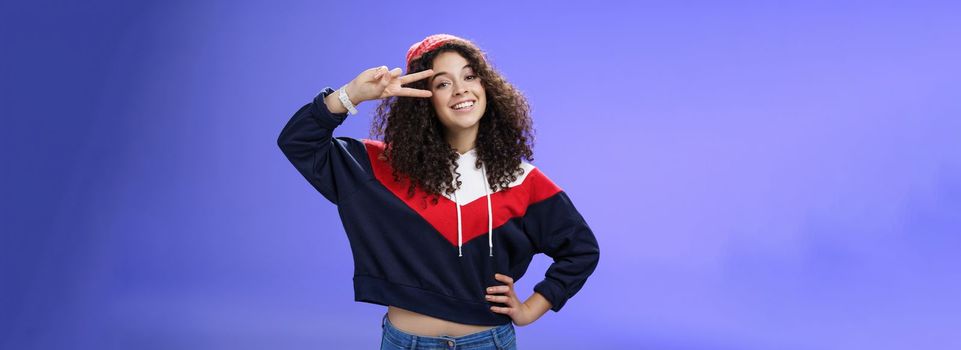 Lifestyle. Attractive feminine woman with curly hairstyle in hat and sweatshirt showing peace or victory sign around eye and smiling carefree having fun playing in yard with snow over blue background.