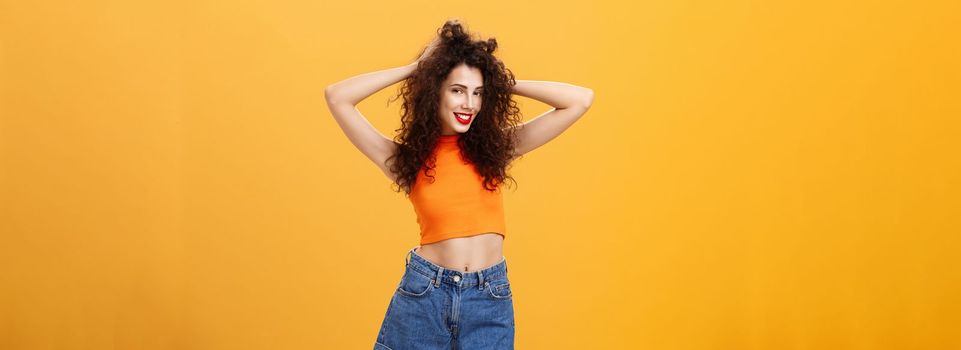 Woman posing for boyfriend. being pleased with new look after visiting hairdresser touching curly hairstyle flirty and carefree smiling sensualy standing over orange background in cropped top.