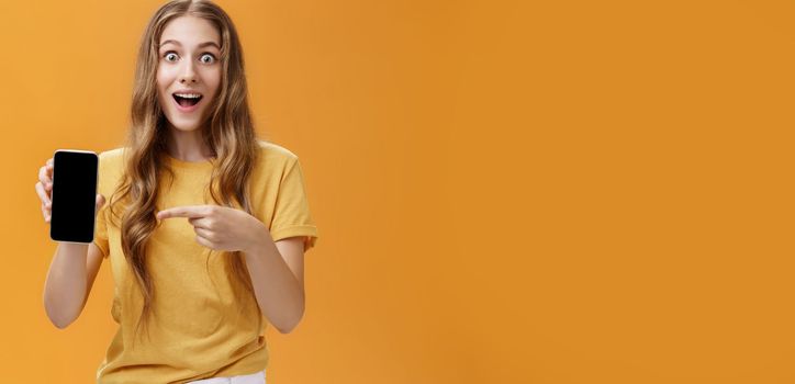 Girl surprised with awesome smartphone features. Portrait of delighted and amazed cute european slender woman with wavy natural hairstyle showing cellphone pointing at gadget screen at app. Copy space