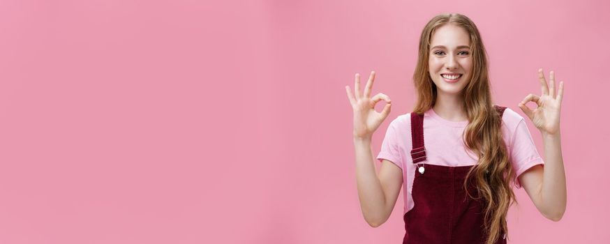 No problem, everything okay. Portrait of pleasant and kind pretty young female in dungarees showing ok or perfect gesture with both hands smiling broadly with friendly gaze at camera over pink wall. Emotions and body language concept