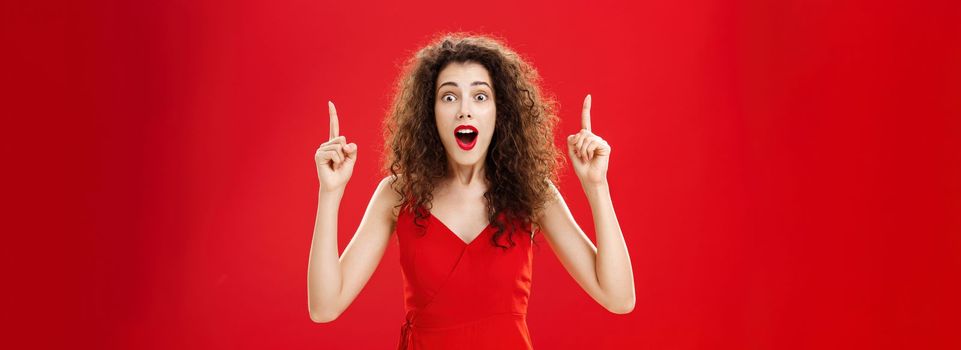 Woman stunned and impressed of huge sales. on clothes gasping and dropping jaw from amazement standing over red background in evening dress with make-up and curly hairstyle pointing up with raised arms.