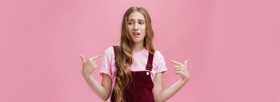 Self-assured arrogant young female student with high ego standing cocky and brag about herself pointing at her with cool snobbish look looking away to right with contempt posing over pink wall. Body language and emotions concept